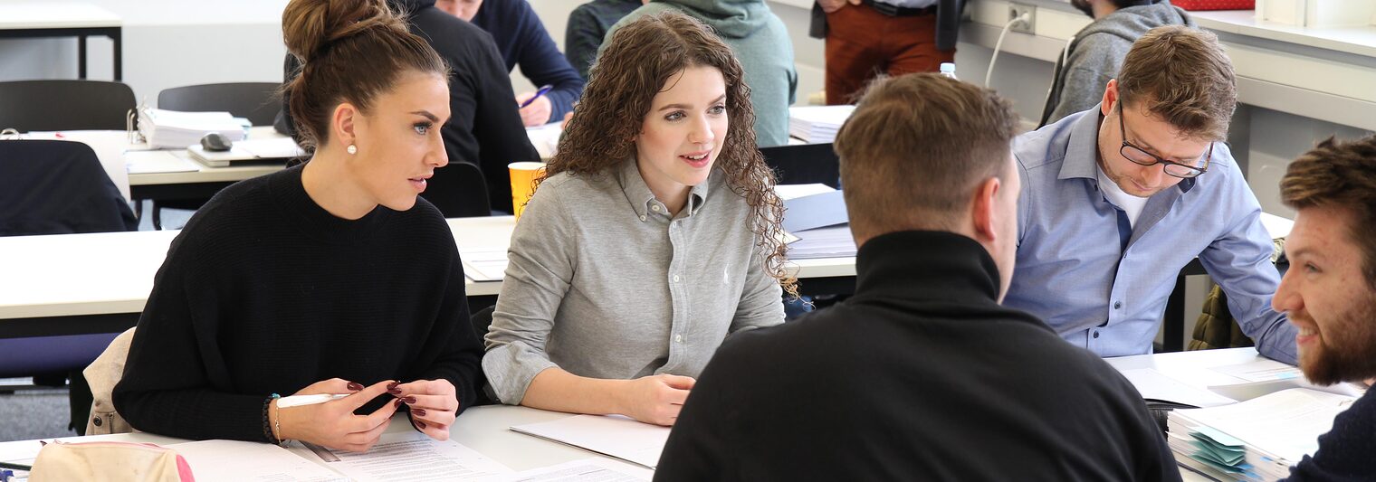 Symbolbild für Weiterbildung, 5 junge Menschen sitzen zusammen an einem Tisch in einem Klassenzimmer