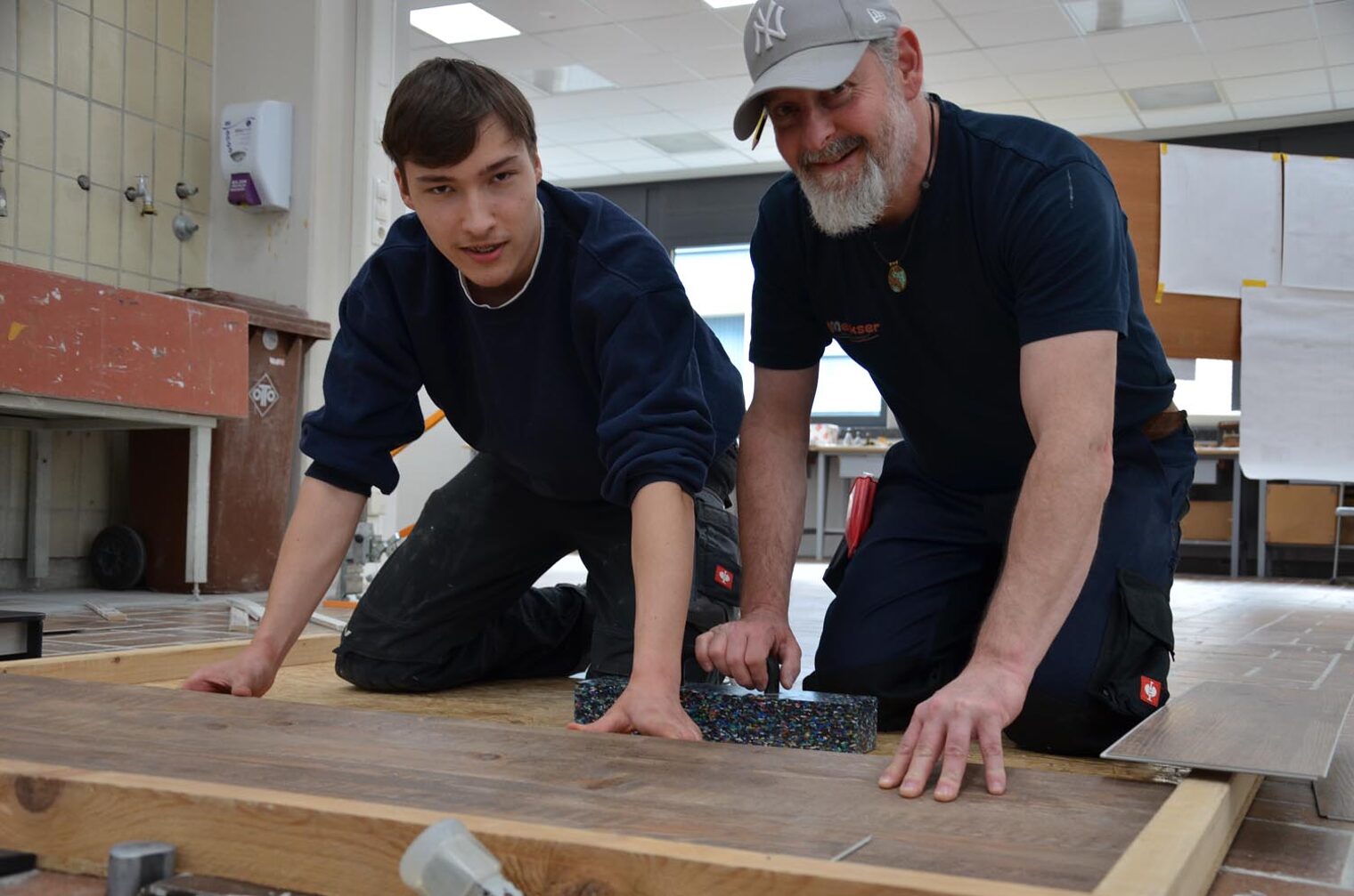 Peter Meeks und Auszubildender Dominik Bund knien auf dem Boden und zeigen das Verlegen von Klick-Vinyl 