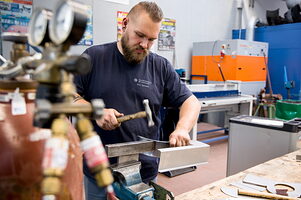 Trainer Max-Ferdinand Gernert bearbeitet in der Karosserie-Werkstatt ein Metallstück mit einem Hammer in einem Schraubstock.